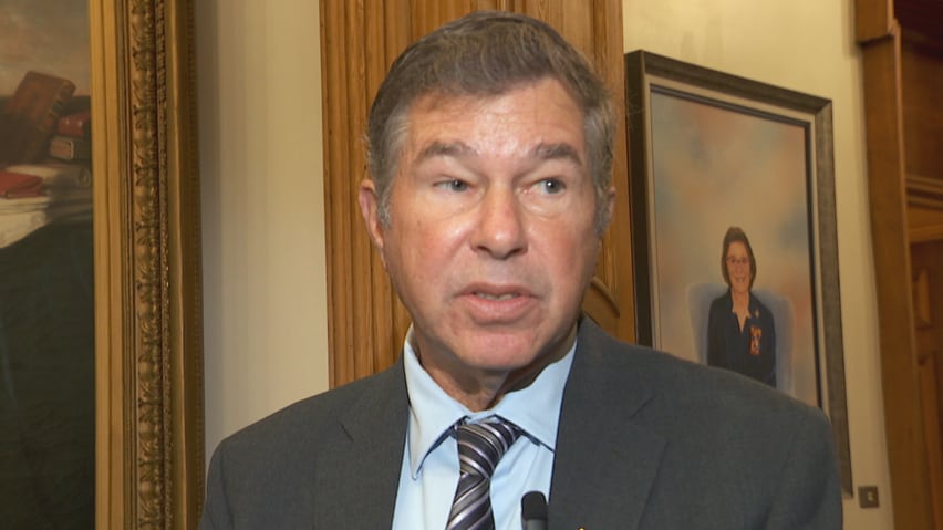 A portrait of a man wearing a suit and a lapel microphone, speaking.