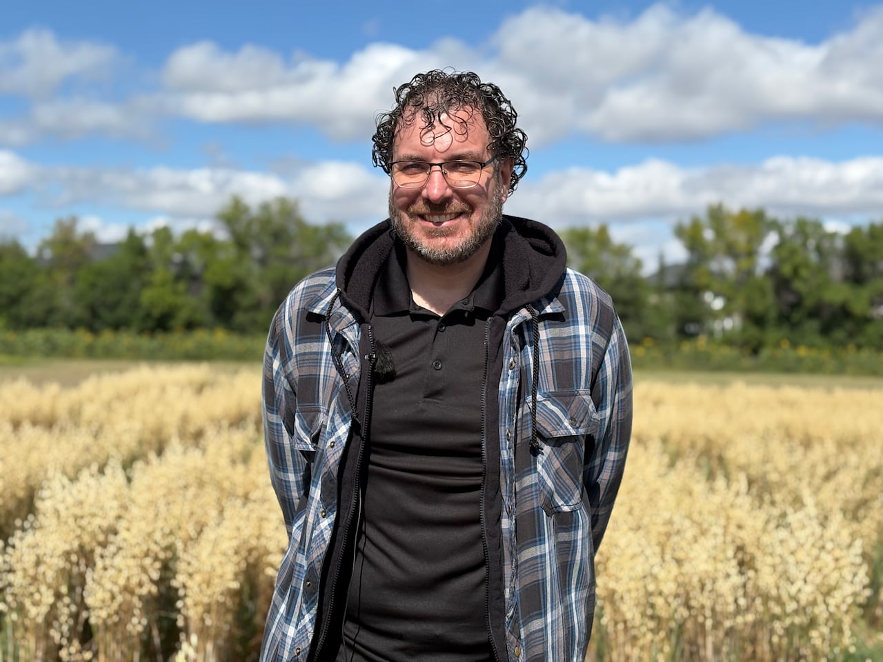A man in glasses, a black shirt and a black and blue plaid jacket stands in a field.