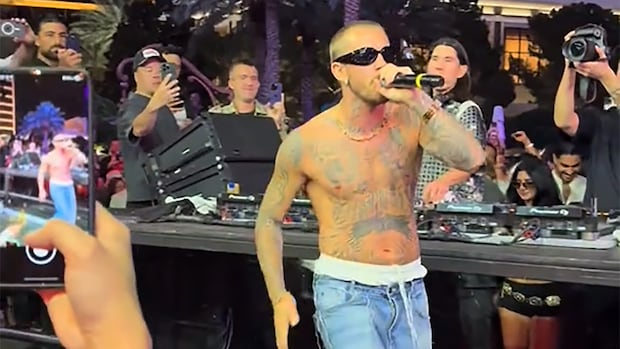 A shirtless, tattooed man in baggy pants and wraparound shades sings into a microphone amidst a crowd. 