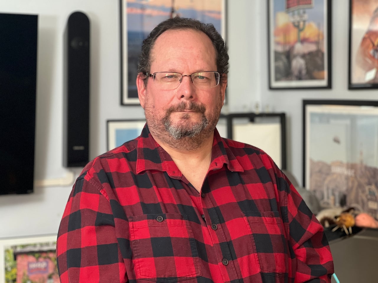 A man wearing a red plaid shirt looks at the camera. He has posters behind him.