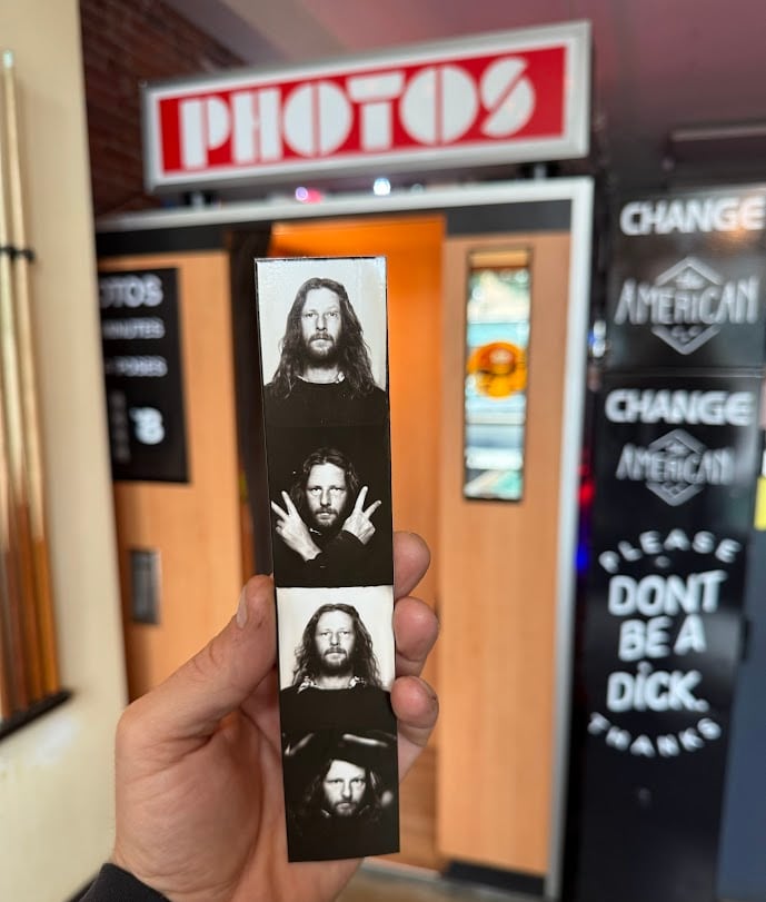 In front of an old-fashioned photo booth, a man's hand holds up a strip of black-and-white portraits of a bearded man with long hair .