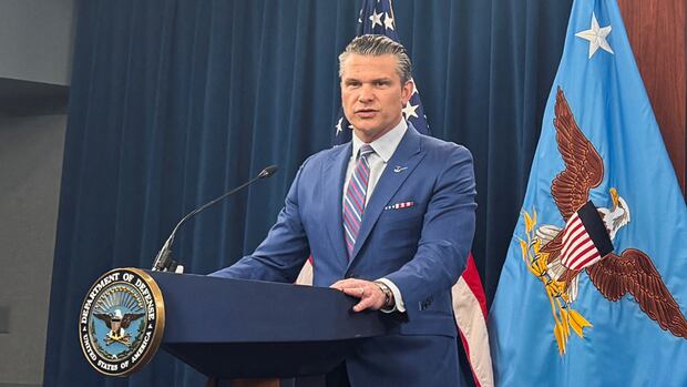 A man in a blue suit speaks at a podium with the Department of Defense seal. An American flag and an eagle emblem flag are in the background.
