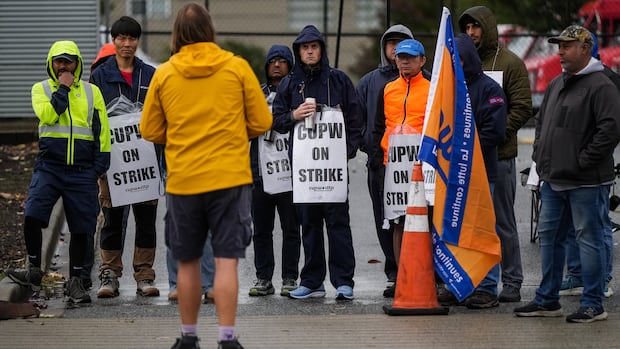 A man in a yellow jacket with his back turned faces a crowd of protesters outside on a rainy day.