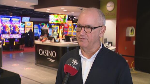 David MacKenzie is the general manager of Red Shores. A balding man with glasses in a white button-down shirt and navy cardigan stands next to a sign saying CASINO, with slot machines in the background.