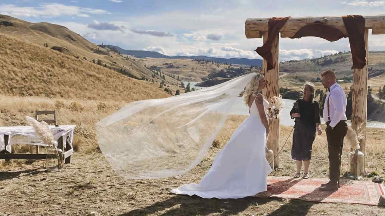 a mates stands astatine an arch with a pistillate holding a microphone betwixt them. the rolling hills of Kamloops are pictured down them