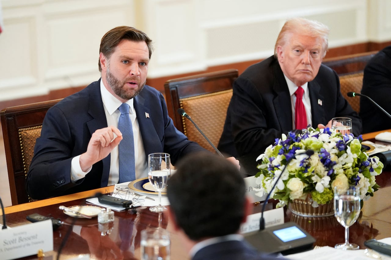 A bearded brownish  haired antheral   successful  a suit   and necktie  speaks portion    seated astatine  a table. Next to him is an older cleanshaven man.