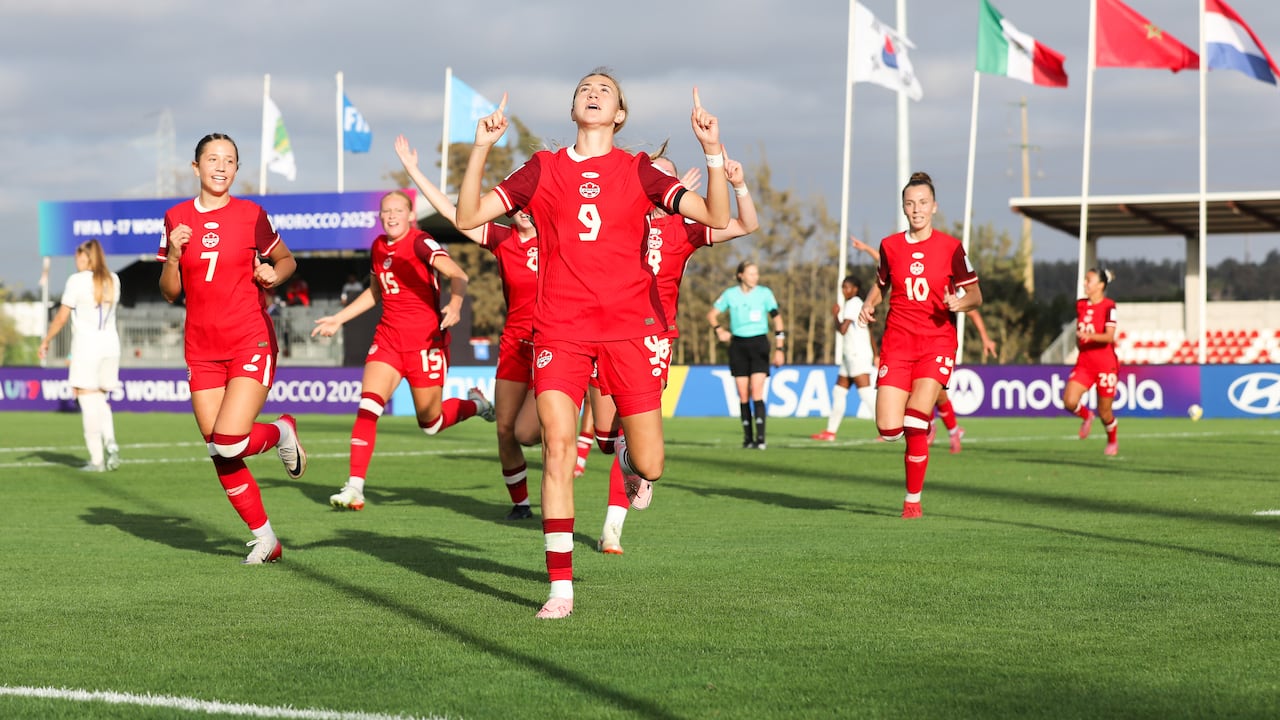 A female soccer player wearing number nine for Canada points both index fingers towards the sky in celebration as her teammates run behind her.