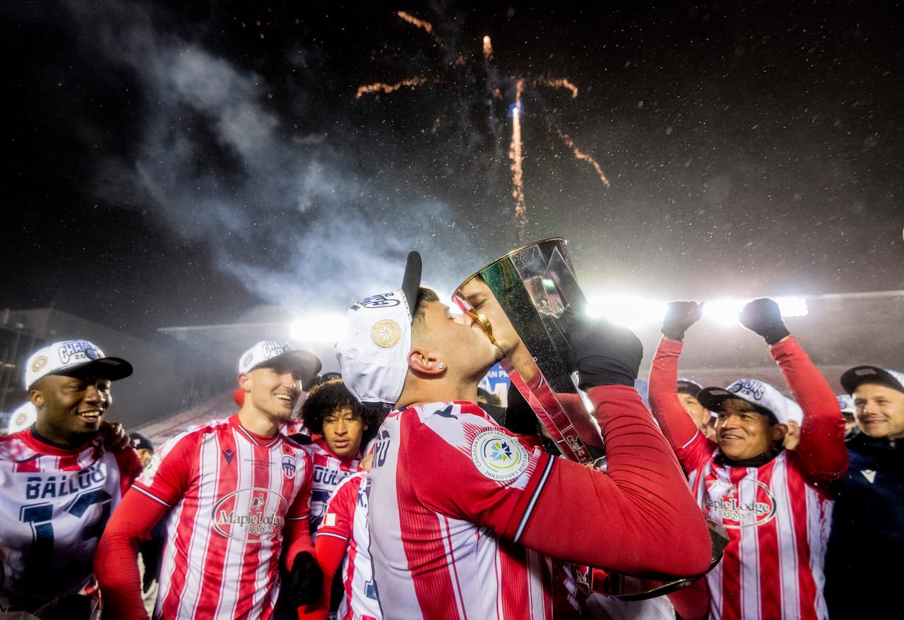 A soccer player kisses a trophy on a snowy night while his teammates gather around him.