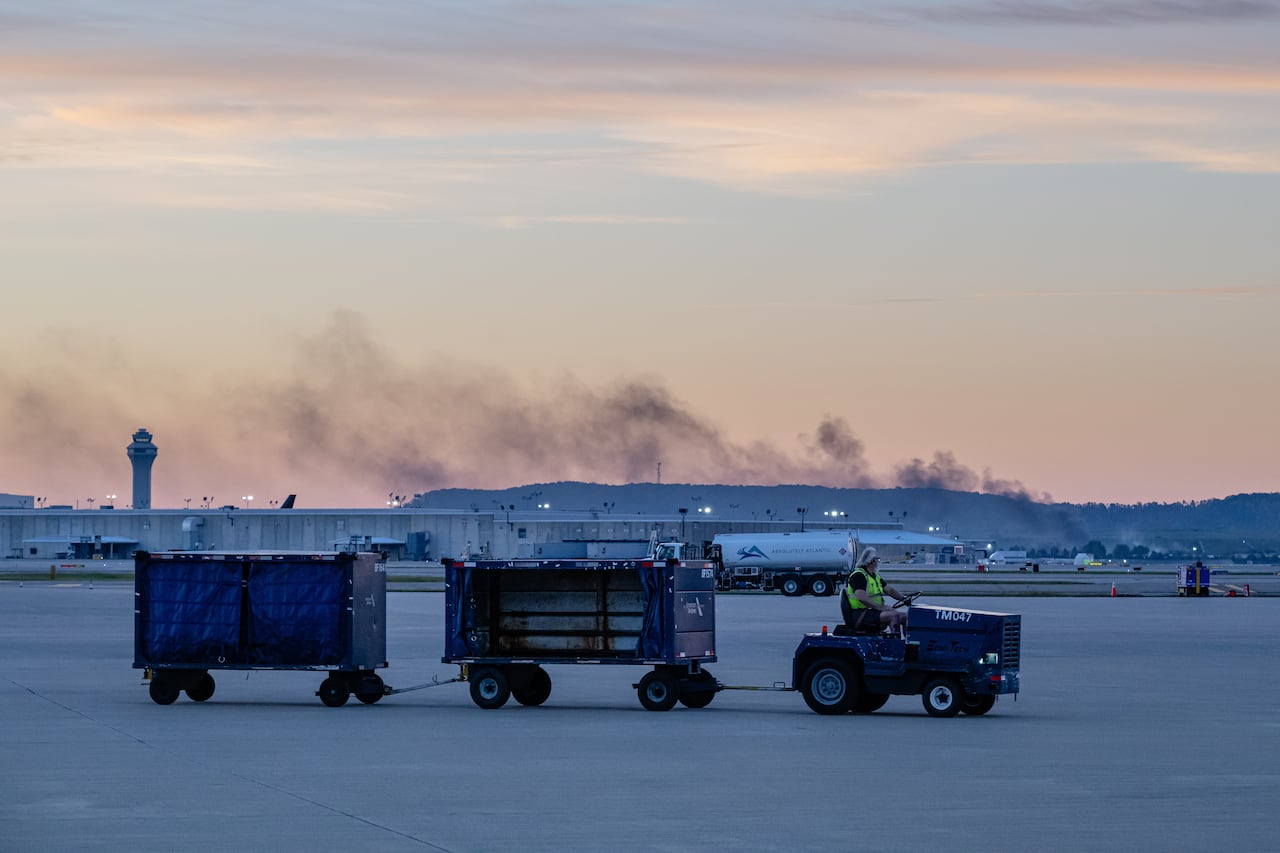 A person drives a tug across an airport tarmac as smoke rises in the background