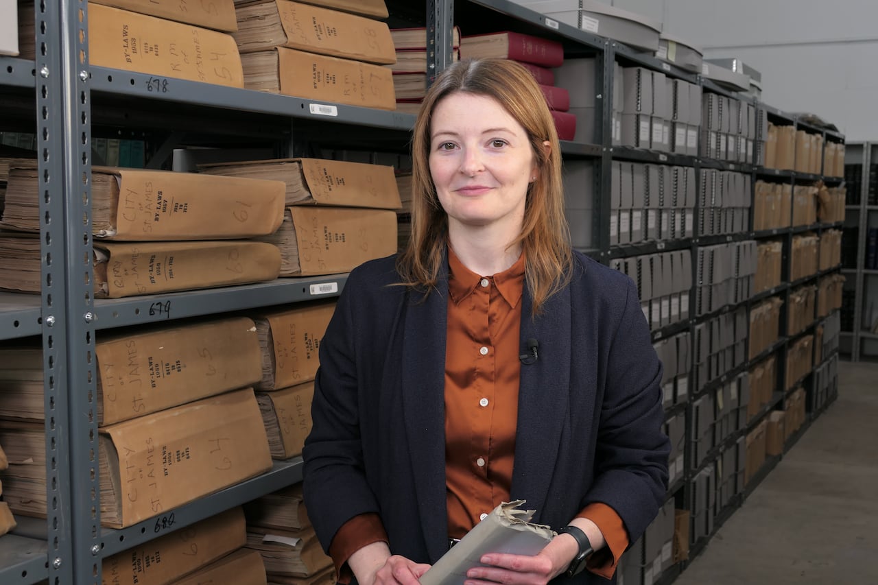 A woman with long red hair stands in a row of documents on bookshelves