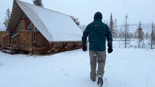 A Parks Canada worker walks towards a cabin, while it is snowing. A Parks Canada worker walks in the snow, towards a cabin.