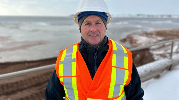 Engineer Brian Thompson has been working on the Souris rock guardian project. Man in white hard hat, orange safety vest and navy winter coat stands in front of a wintry ocean shoreline.