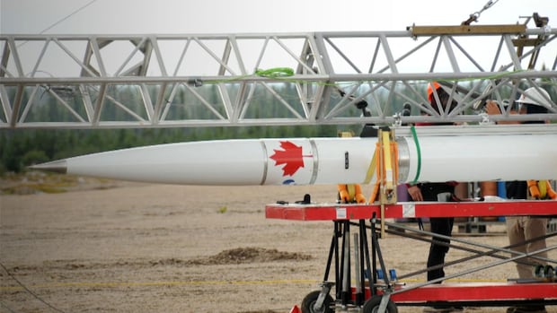 A white rocket with a red maple leaf painted on it is being prepared for its launch from Cree land.