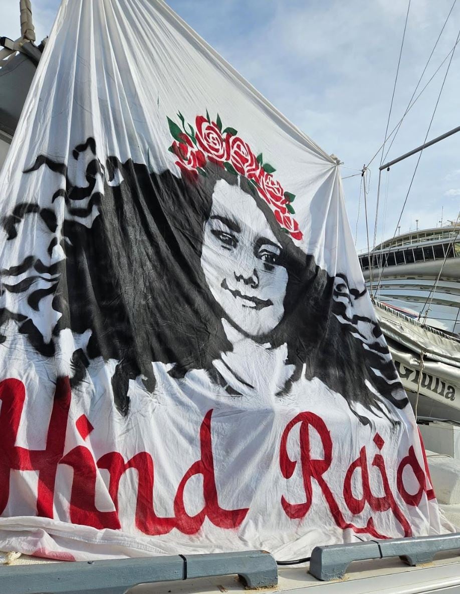A large sail for a boat with a painting of a young girl on it with the name Hind Rajab written on the bottom.