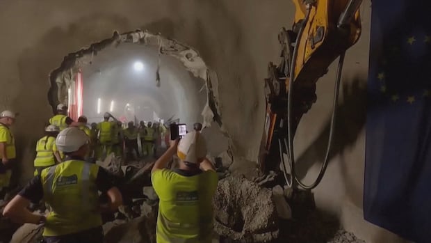 A group of people in helmets and yellow shirts stand next to a hole in rock and an excavator