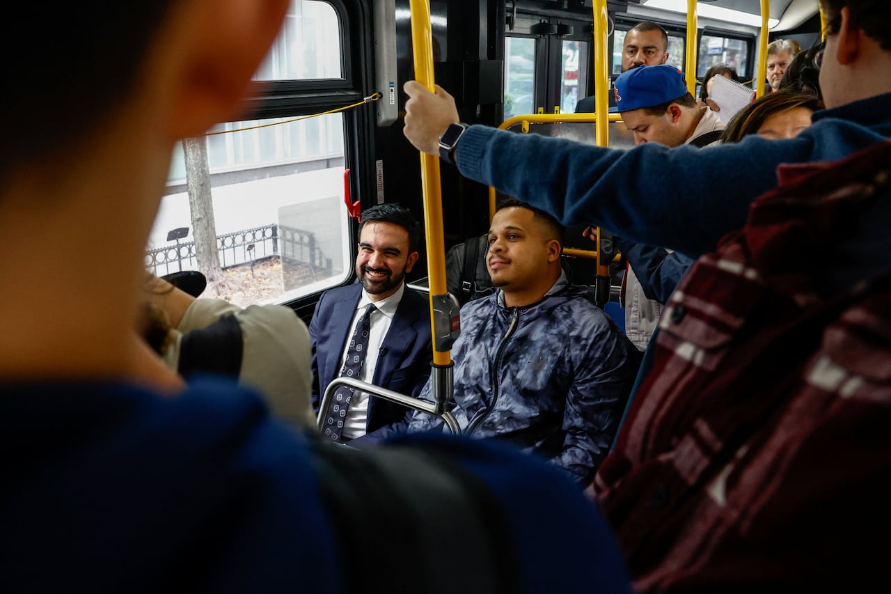 A man in a blue suit sits on the bus, smiling as the passengers look on.