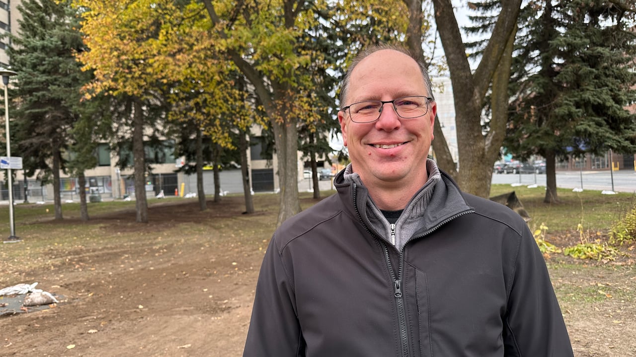 A bald antheral   wearing a grey windbreaker stands against immoderate   bare crushed  with trees successful  the background