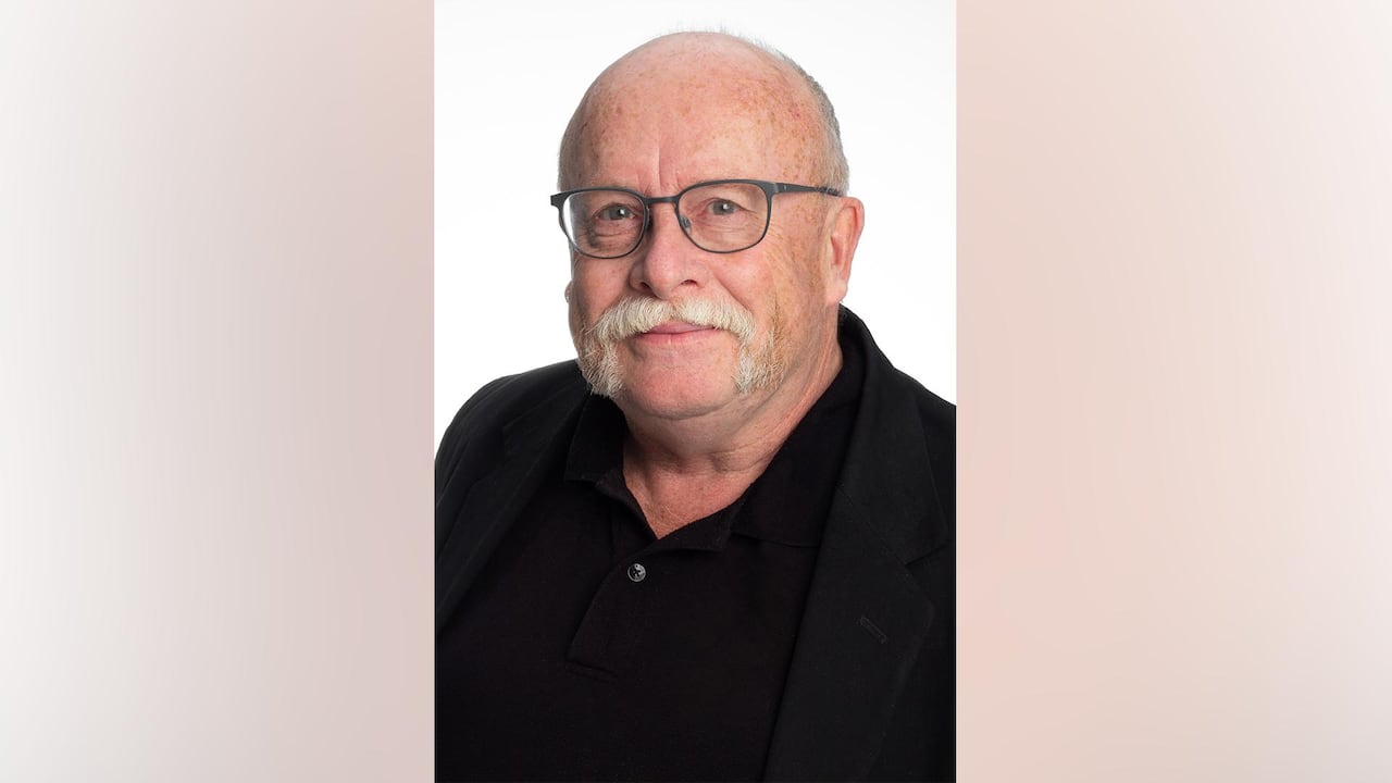 A portrait of an older, mustached man in glasses dressed in a black shirt and blazer.