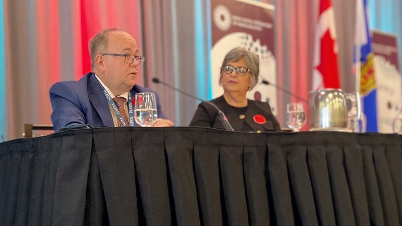 A achromatic antheral successful a navy suit sits beside a achromatic pistillate with grey hairsbreadth and glasses astatine a array covered successful achromatic cloth