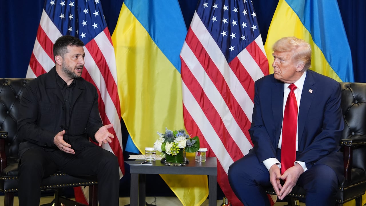 A man in a black suit sits across from a man in a blue suit and red tie, with flags behind them.