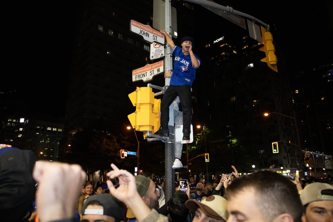 A young man in a Blue Jays jersey scales a street sign above a crowd of Jays fans at a Toronto intersection at night