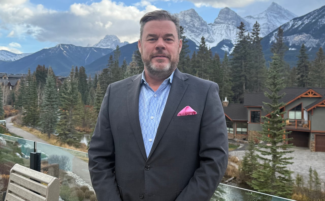 A man in a suit stands on a balcony against a background of trees and mountains.