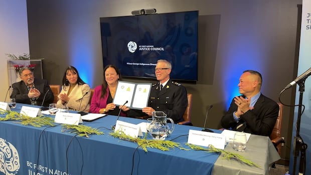 Five smiling people sit at a blue conference table in front of microphones. The two in the middle hold up a an agreement they have signed.