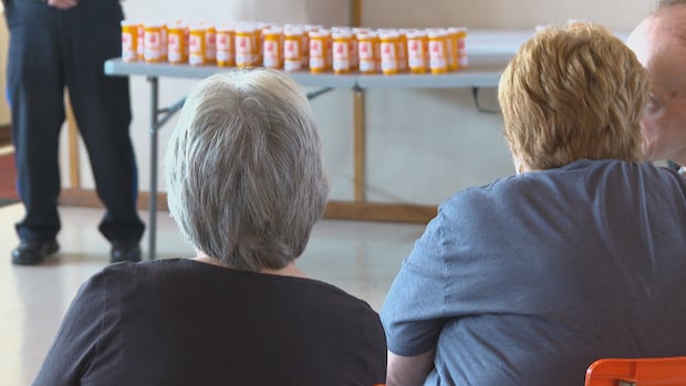 Two women, both seniors, can be seen from behind. In front of them is a table filled with pill bottles.