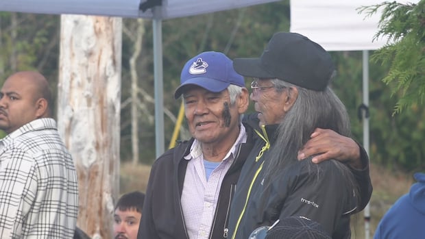 Thomas Rush, wearing a Vancouver Canucks hat, attended the residential school. He said he wanted to witness its demolition as a way to provide healing. Two men grasp each other's shoulders.