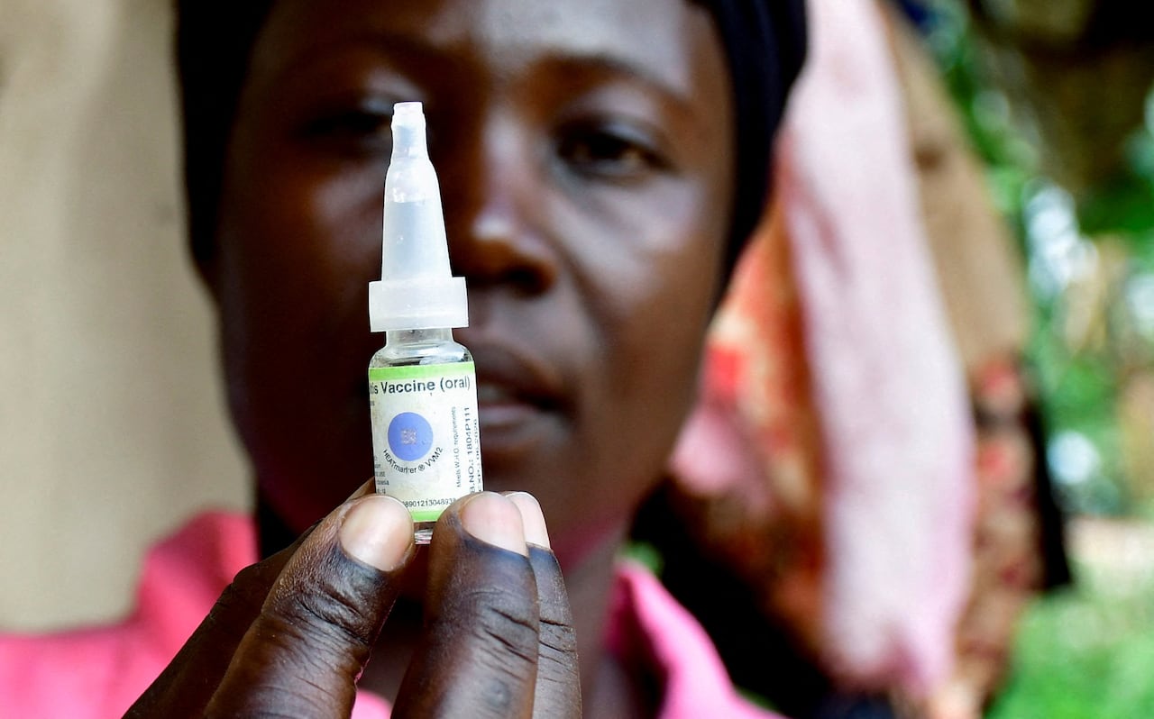 a womans face is blurred in the background, and in front of her face in focus she holds a vial of polio vaccine