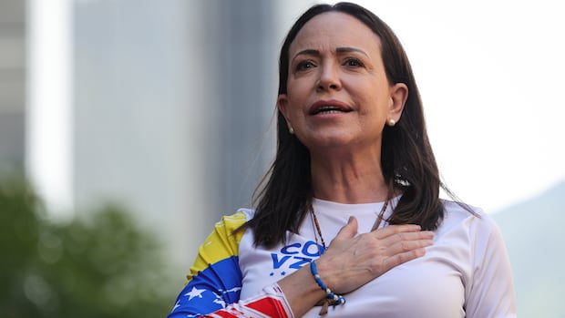 A woman with brown hair just past her shoulders puts her hand over her chest while standing in an outdoor setting.