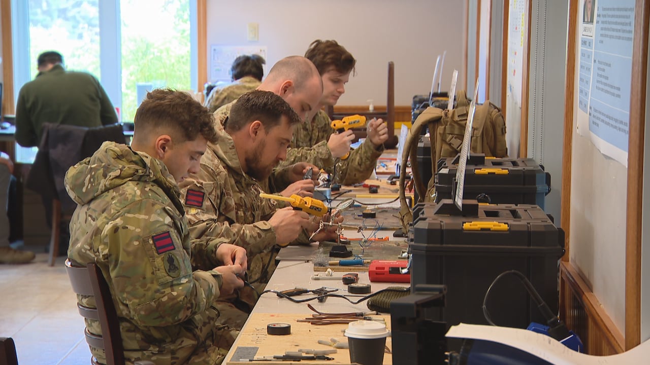Four men wearing military uniforms sit in a row using yellow glue guns. 