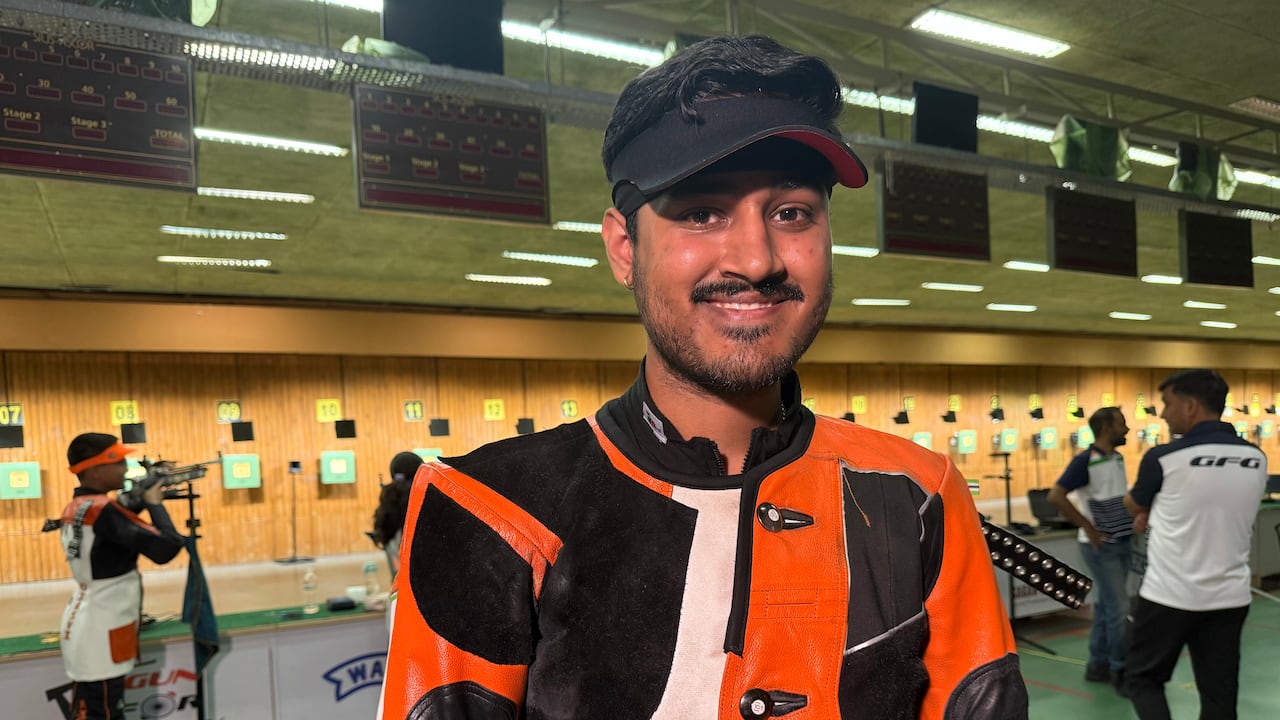 一名身穿黑、白、橙色夾克、戴著黑色面罩的年輕人站在射擊場前。