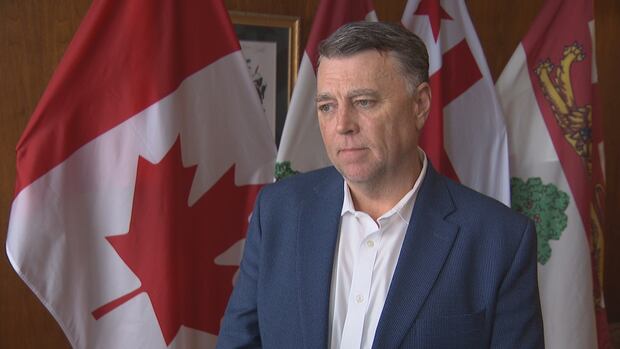 Man in a blue blazer and white shirt stands solemnly in front of Canadian flags. The setting conveys a serious, official tone.