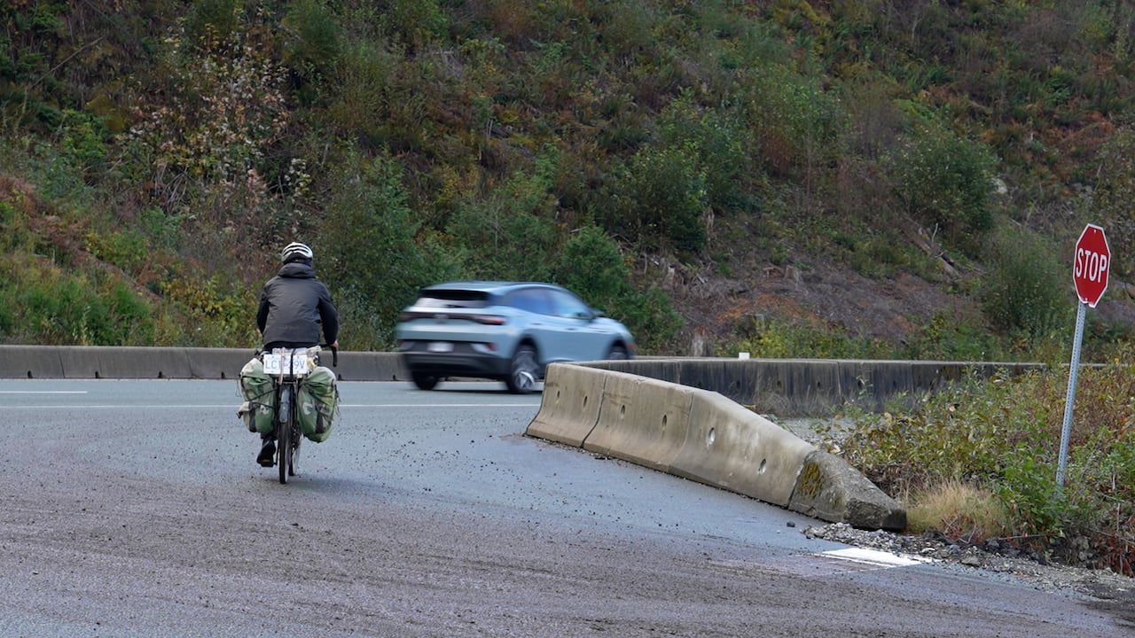 A man rides a bicycle on the left side of the frame as vehicles pass on the road.