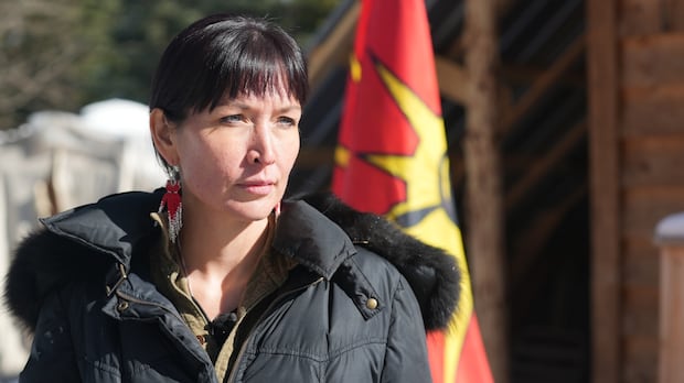 Sleydo', also known as Molly Wickham, is a Wing Chief of Cas Yikh, a house group of the Gidimt'en Clan of the Wet'suwet'en Nation in B.C. A dark-haired woman stands in front of a warrior flag