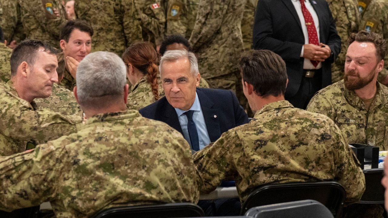 A group of soldiers in camouflage uniforms sit around a table with a man in a suit. The setting is serious yet collaborative, indicating a discussion.