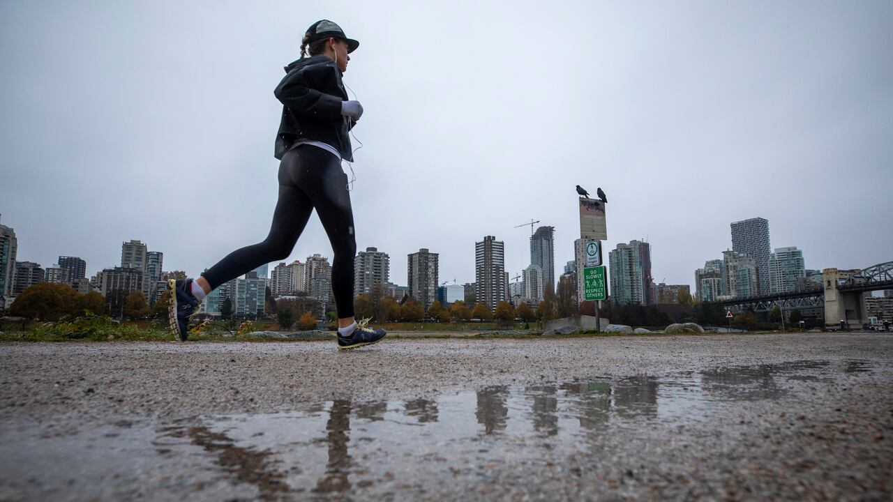A woman runs along a muddy trail, with a city skyline visible behind her, on a rainy day.