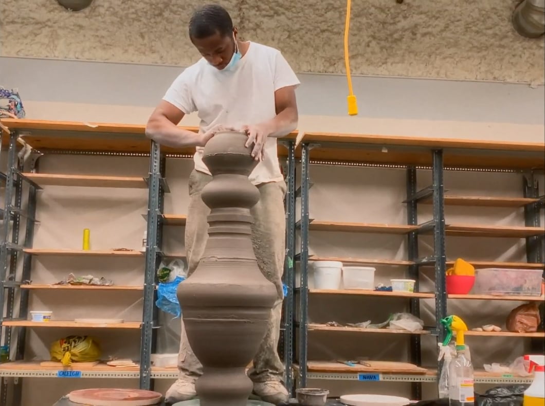 A man, wearing khaki pants and a white t-shirt, stands on a table inside an art studio while shaping a large clay vessel.