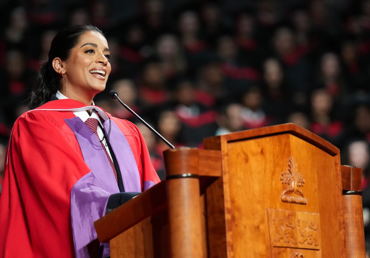 A woman in a red and purple graduation gown speaks into a microphone on a wooden podium.