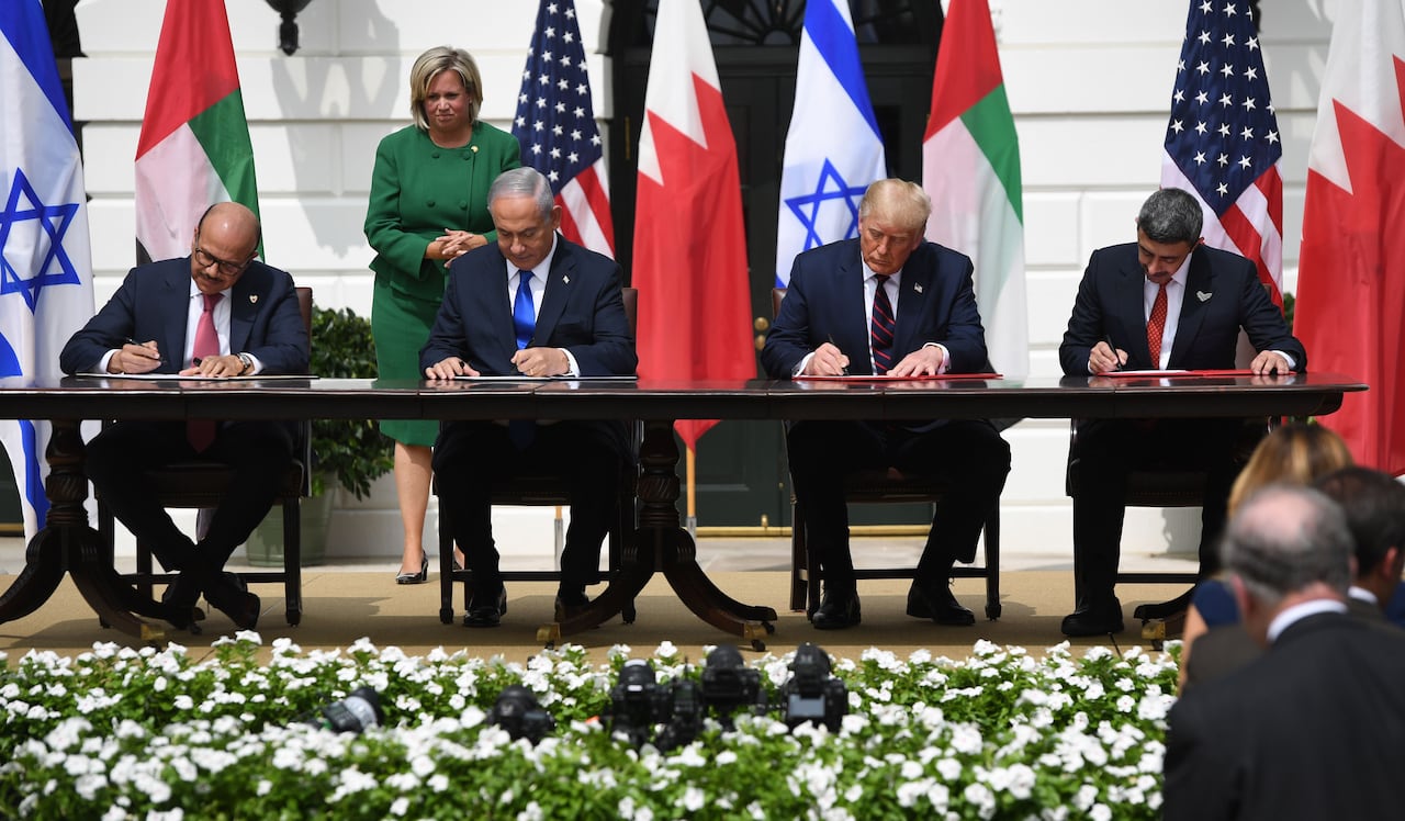 Four men in black suits and ties sit at a table, each signing a document placed in front of them. Colorful flags are visible behind them.