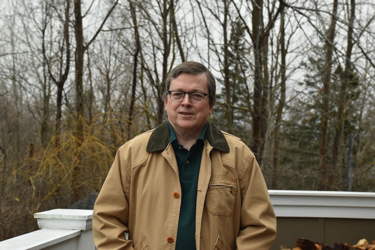 A man in a camel coloured trench stands against a backdrop of trees.