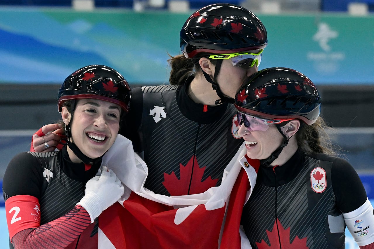 Three speed skaters hug while holding a large Canadian flag.