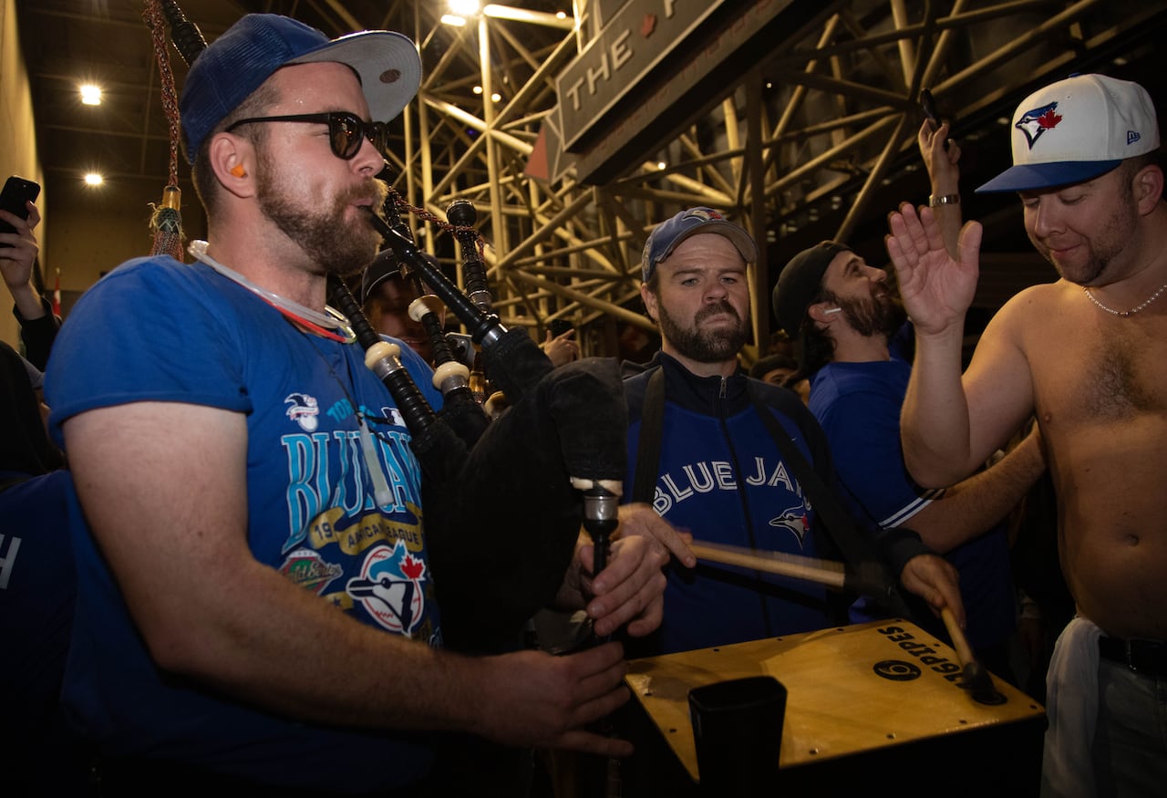 A bagpiper in a Jays shirt plays with a percussionist in a Jays jersey amidst a crowd of fans. One shirtless fan in a Jays cap dances beside them. They are on a city street at night