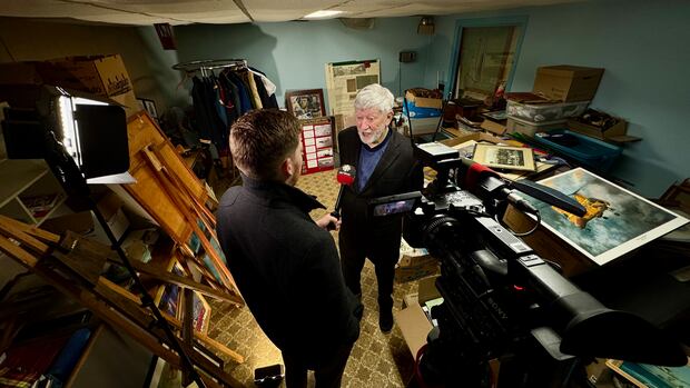 George Dalton being interviewed by reporter Connor Lamont at 200 Summerside Wing club. A man with white hair and beard is interviewed by a younger man in a room packed with boxes. paintings, photographs and military uniforms.