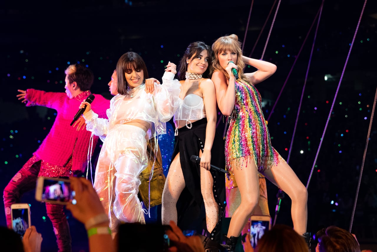 Three women with mics in their hands dressed in sparkly performance outfits stand together on a concert stage.