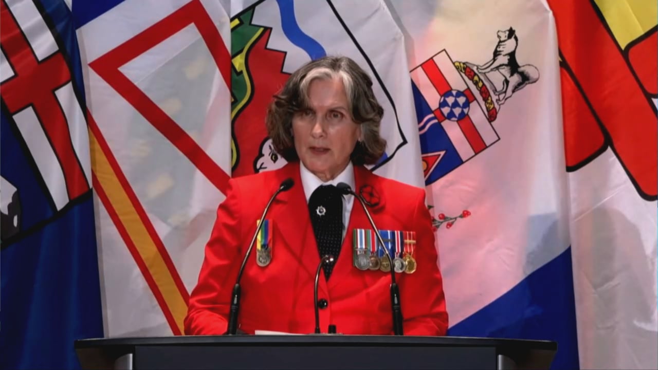 Uma mulher vestida de vermelho e com medalhas militares fala em um pódio.