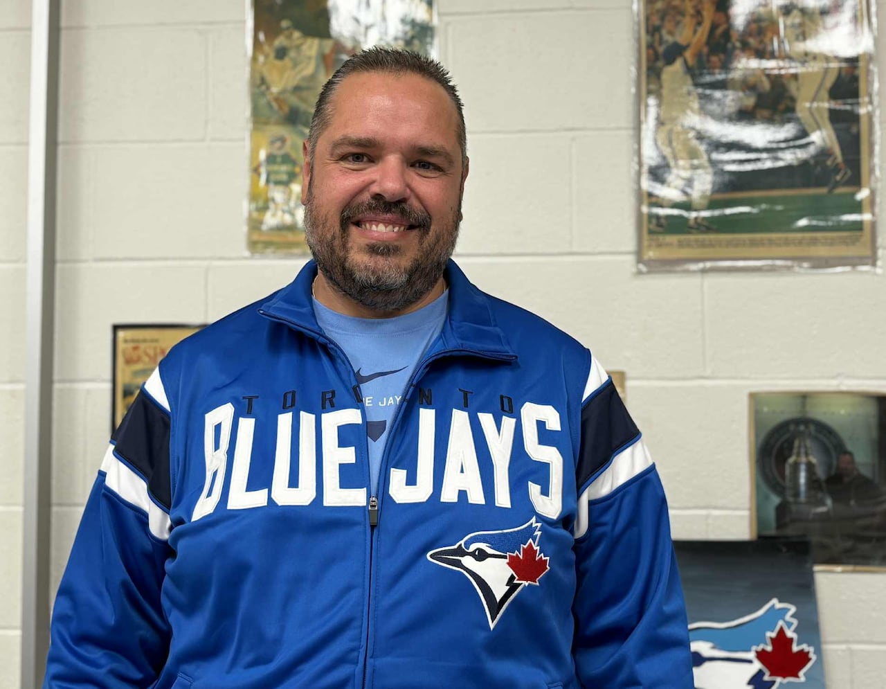 A man in a Toronto Blue Jays jacket stands in front of a wall with sports memorabilia