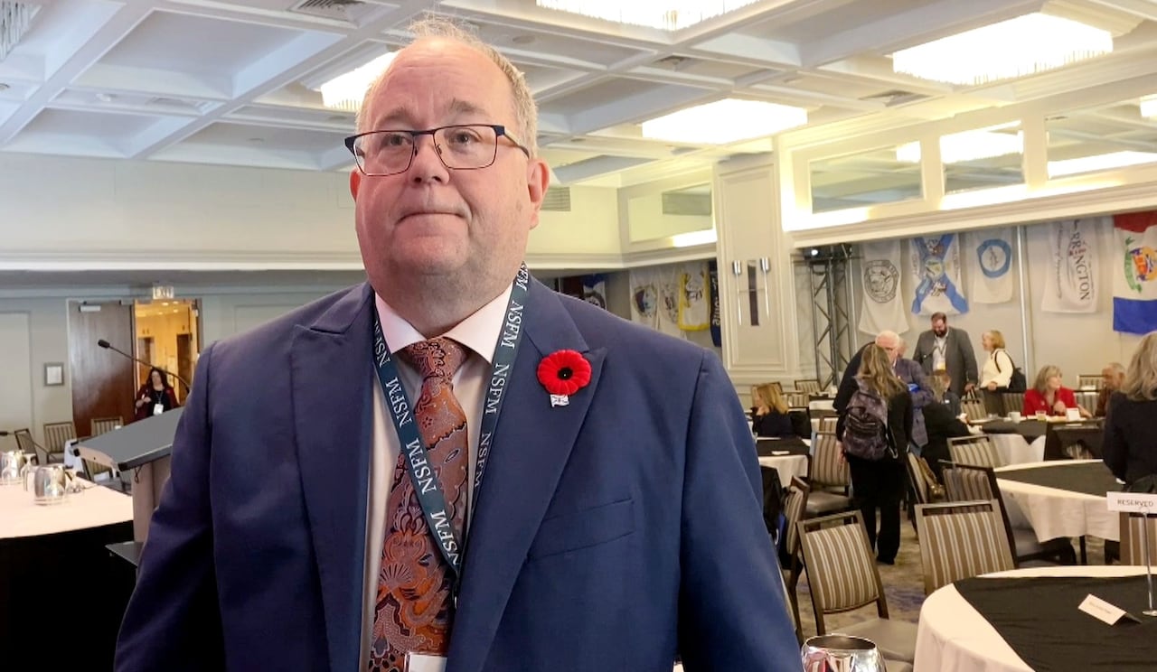 A achromatic antheral successful a navy suit and reddish poppy connected his thorax stands successful a edifice ballroom, with tables down him and radical leaving the room