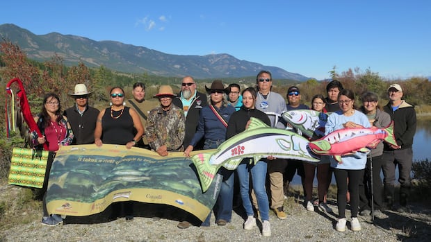 Mark Thomas, in back wearing blue medallion, with other supporters of the Bringing the Salmon Home Initiative at an announcement in September 2025. 16 people hold a banner shaped like a salmon.